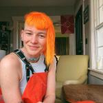 Mace sits wearing an orange overalls jumpsuit with matching bright orange hair.